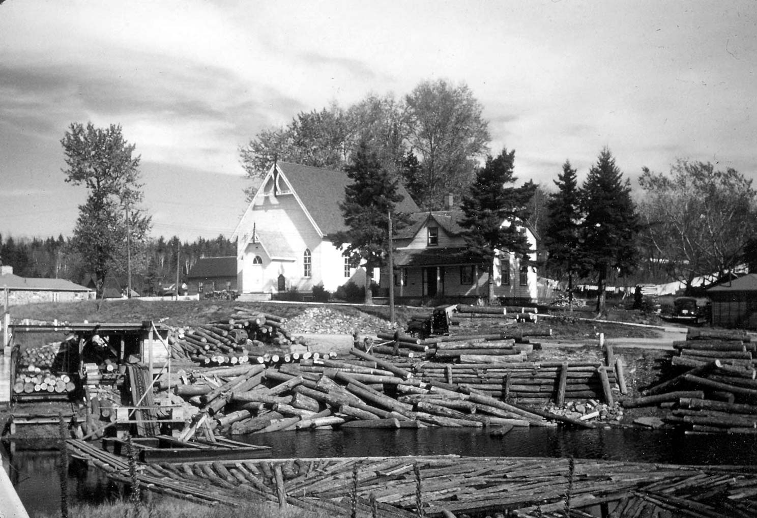 Lots of logs piled up in the river in the foreground of the picture. The Church is in the background and is a white building with a peaked roof.